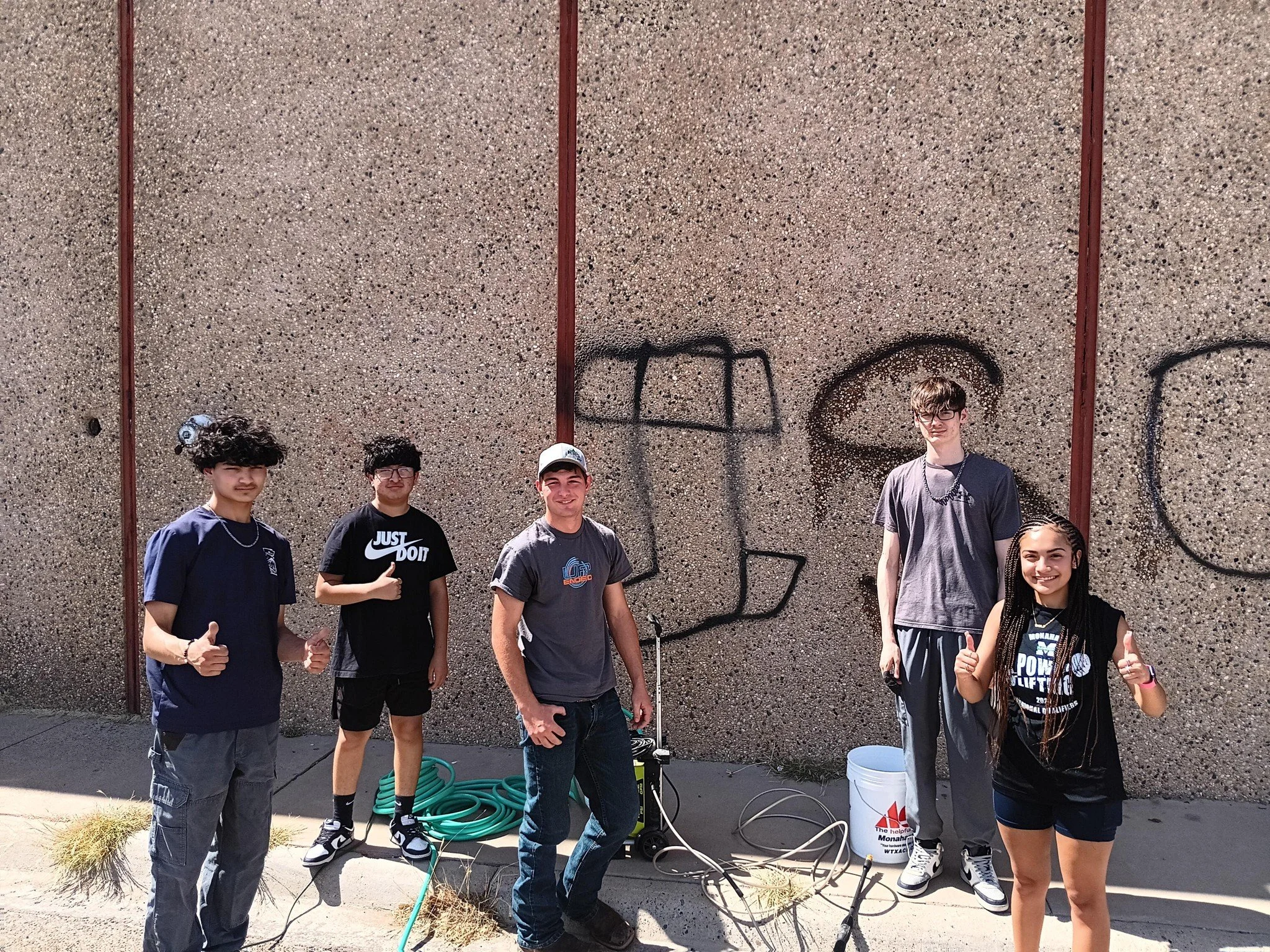 Five teenagers standing outdoors in front of a graffitied wall, with some spray paint cans and a bucket on the ground, giving thumbs up and smiling.
