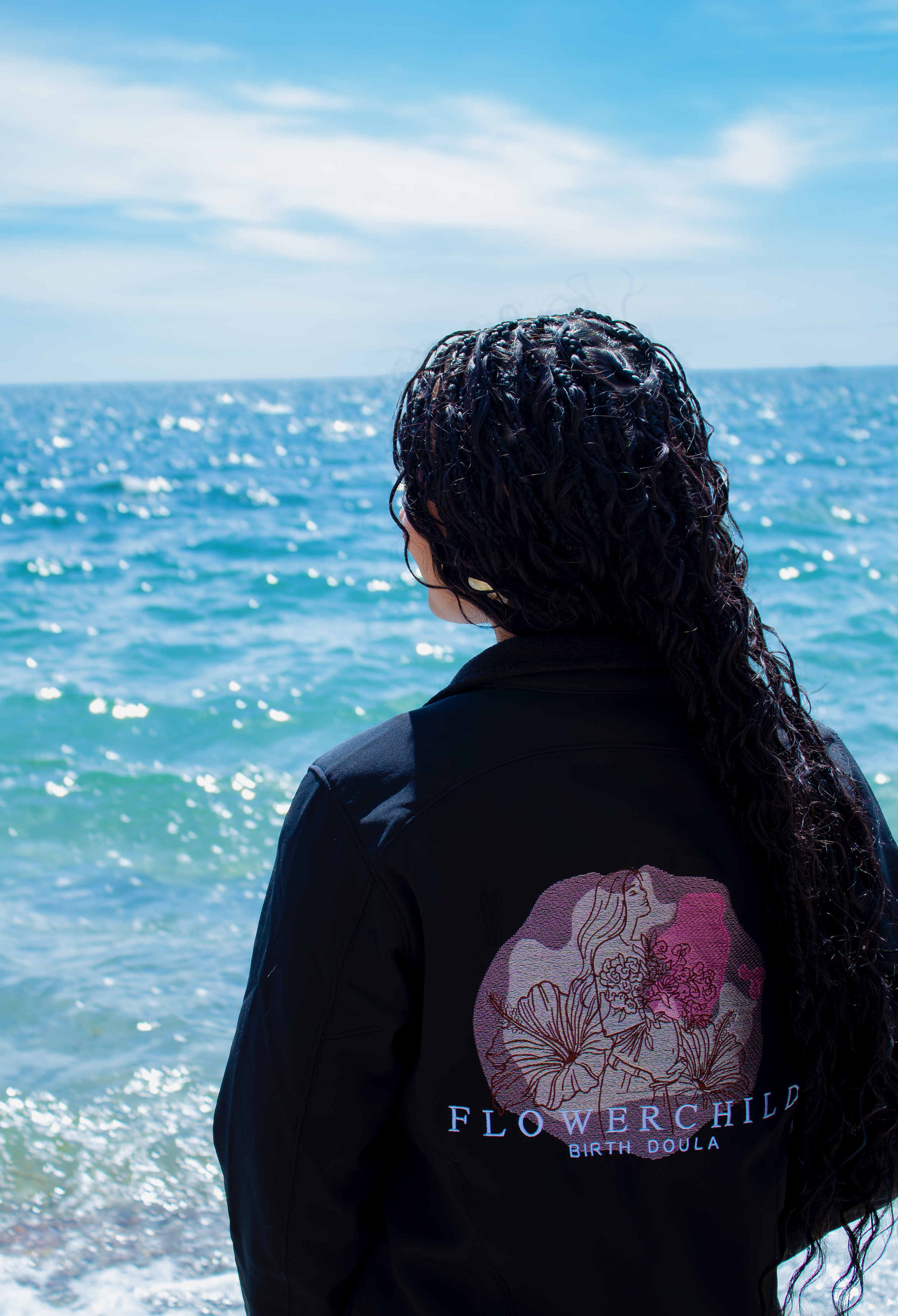 Person with curly hair standing by the ocean, facing away from the camera, wearing a black jacket with a floral design and the words 'FLOWER CHILD BIRTH DOULA' on the back.