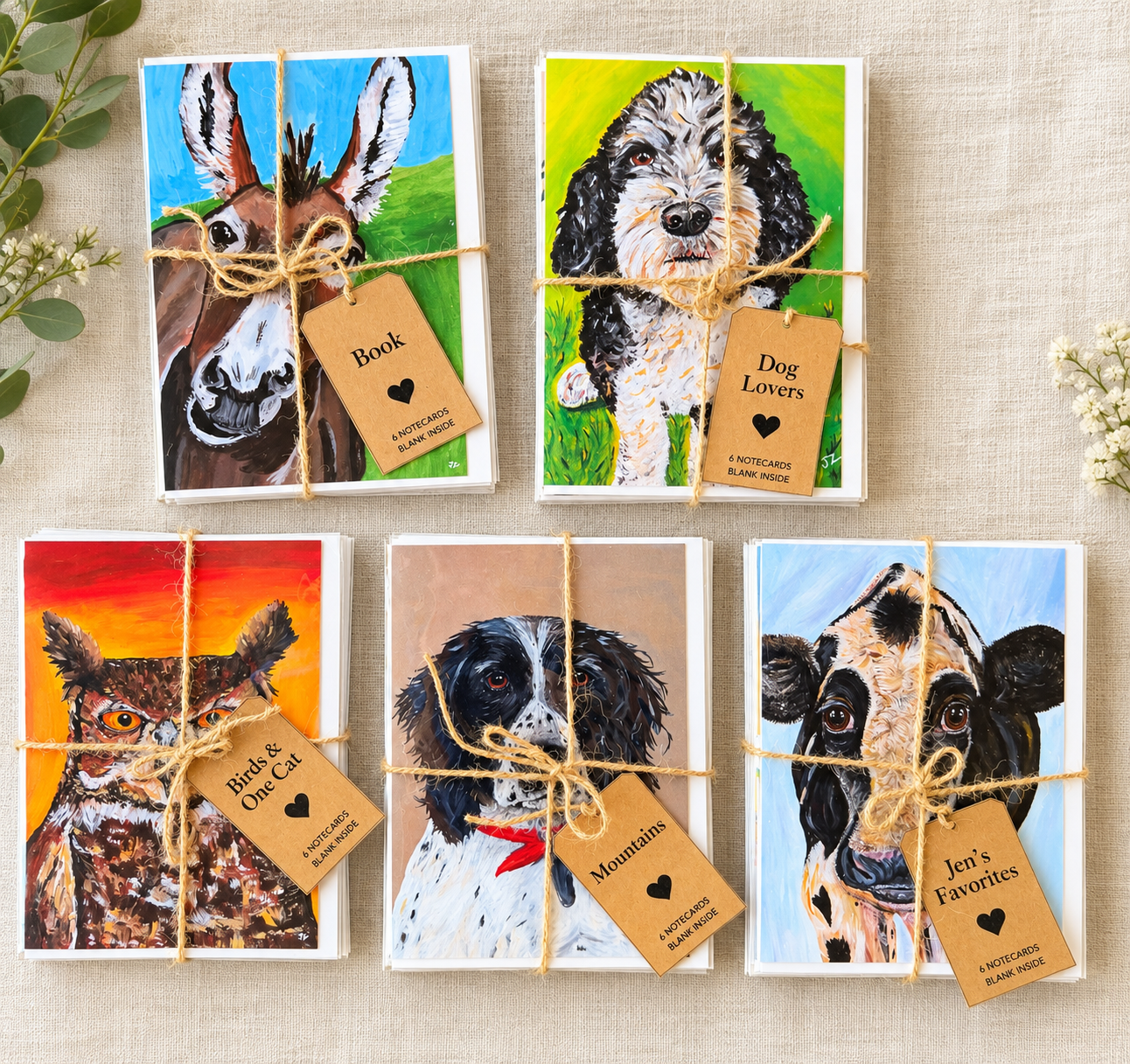 Sets of notecards featuring painted dog and bird portraits, tied with twine, with tags indicating themes like 'Book,' 'Dog Lovers,' 'Birds & One Cat,' 'Mountains,' and 'Jen's Favorites.'