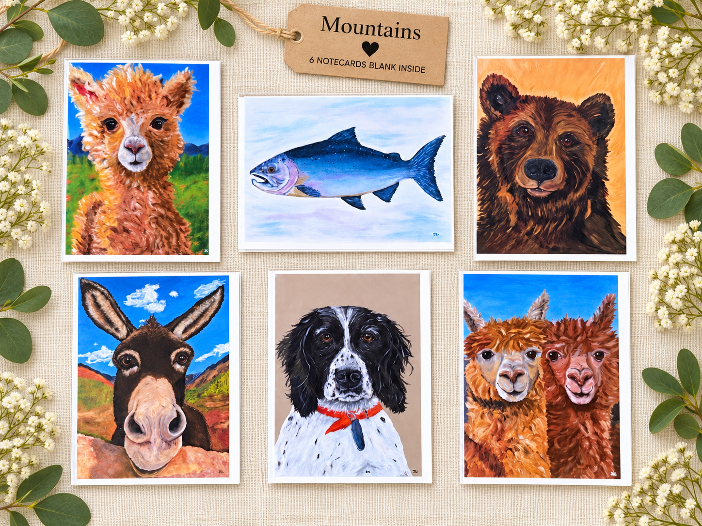 Collection of six painted animal portraits and a painting of a fish, arranged on a beige fabric background with greenery and a wooden tag labeled 'Mountains', indicating these are mountain-themed animal paintings for a set of six blank notecards.