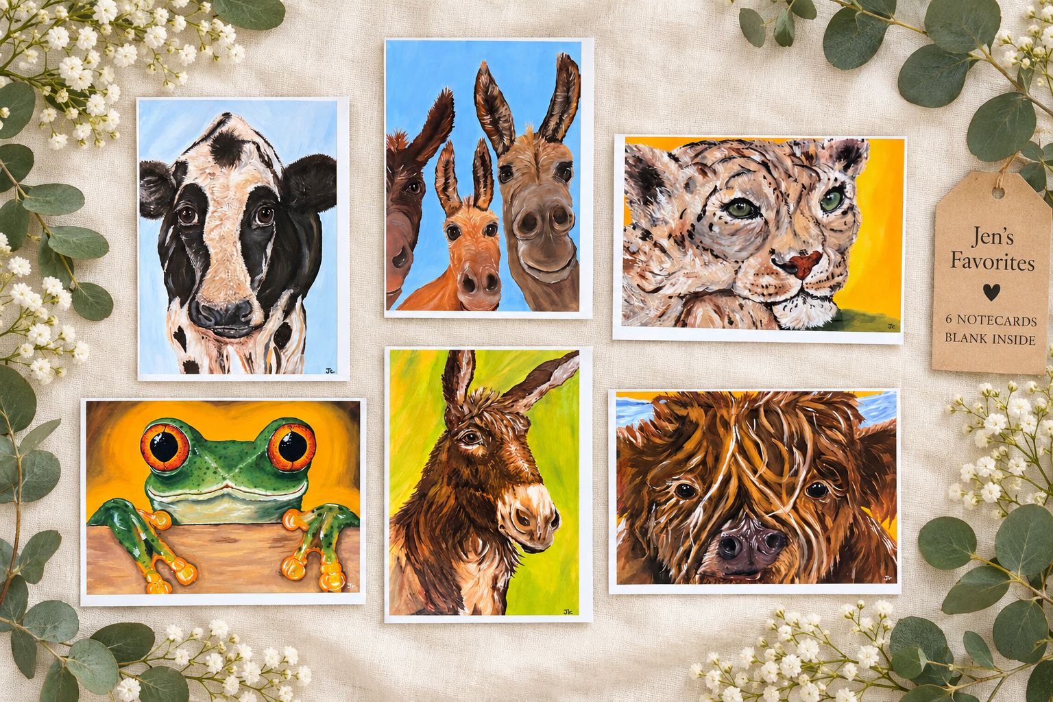 A display of six colorful animal paintings on a beige fabric background, surrounded by white flowers and green leaves, with a tag on the right that says "Jen's Favorites," mentioning six notecards inside.