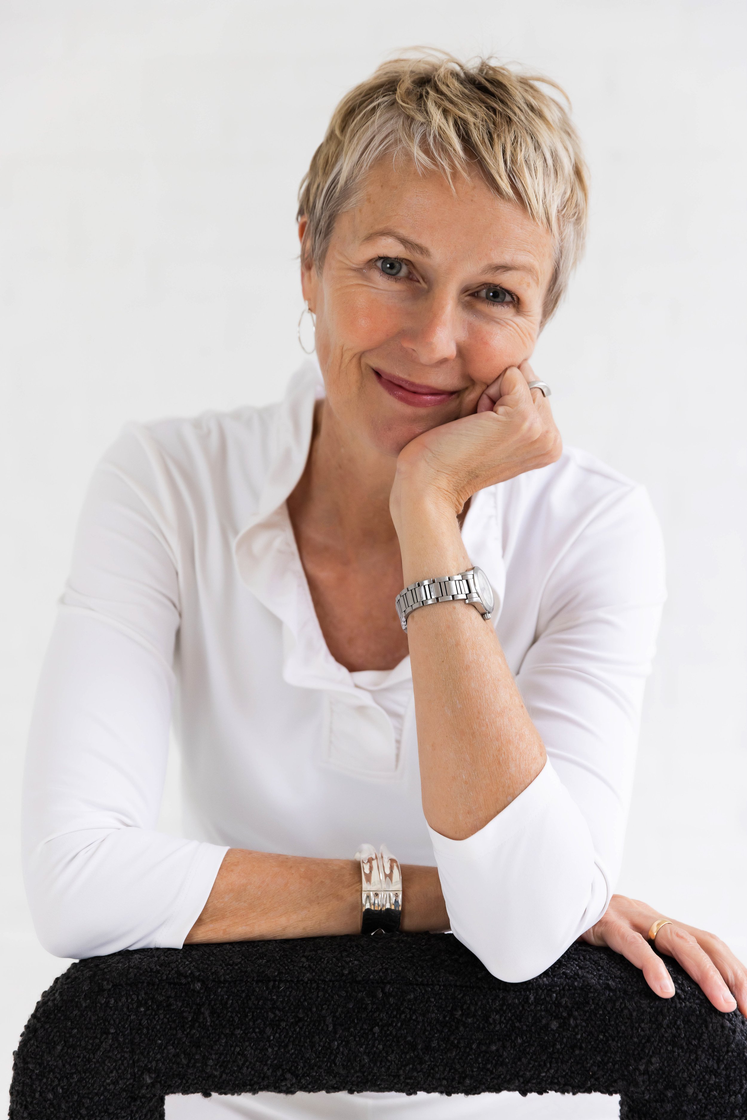 A woman with short blonde hair, light skin, and wearing a white blouse, seated with her left arm resting on a table covered with black fabric, smiling gently at the camera.