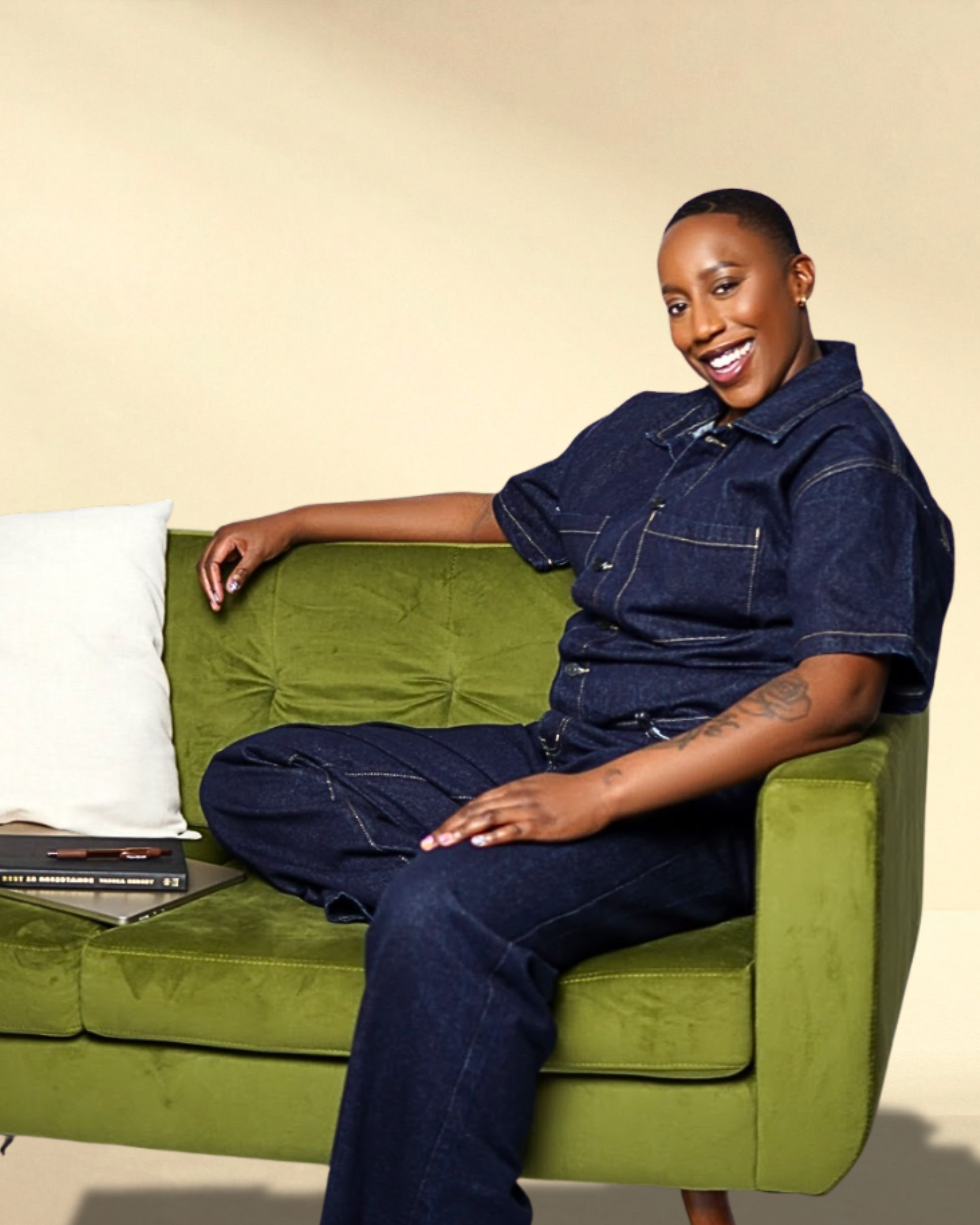 Say More Therapy based in Maryland. Therapist Ramelle Liverpool with short hair and a tattoo on her arm, wearing a dark denim jumpsuit, sitting on a green couch with a white pillow and a closed book, smiling at the camera.