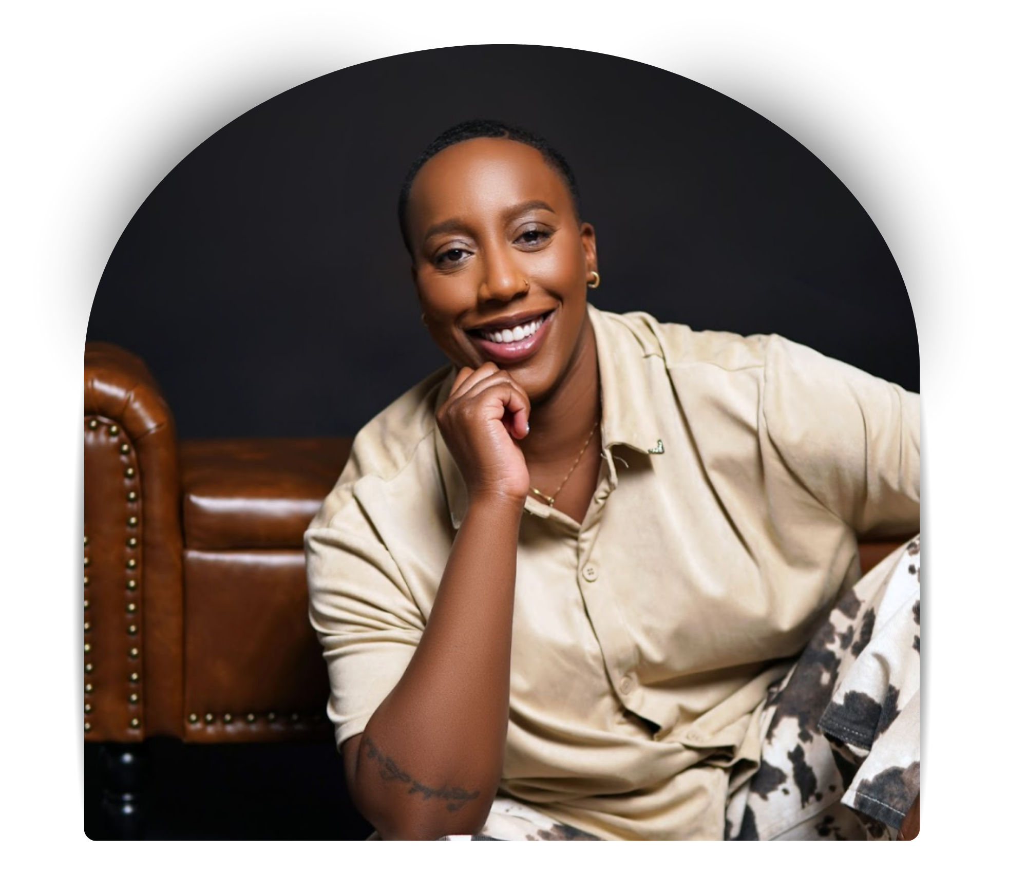 Say More Therapy based in Maryland. Therapist Ramelle Liverpool with short hair and a tattoo on her left forearm, sitting on a leather couch against a dark background, wearing a beige shirt and light-colored pants.