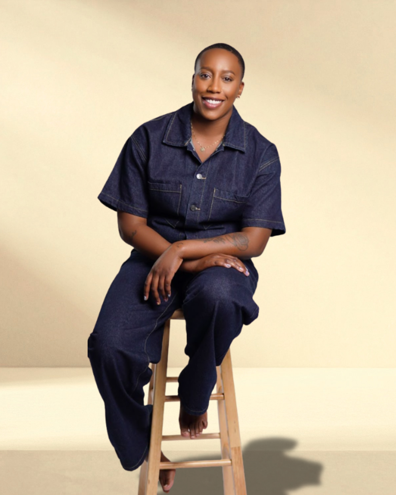 Say More Therapy based in Maryland. Therapist Ramelle Liverpool with short hair wearing a denim jumpsuit, sitting on a wooden stepladder, smiling, against a plain beige background.