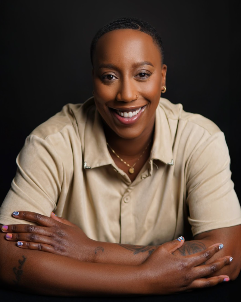 Say More Therapy based in Maryland. Therapist Ramelle Liverpool with short hair and small earrings, wearing a beige shirt, gold necklace, and displaying tattoos on her crossed arms against a black background.