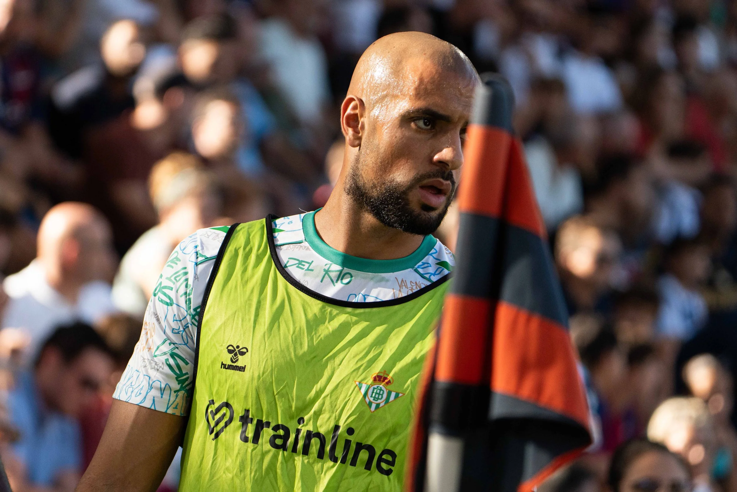 Un futbolista con camiseta de entrenamiento y un peto amarillo, en un partido, con mucha gente en las gradas.