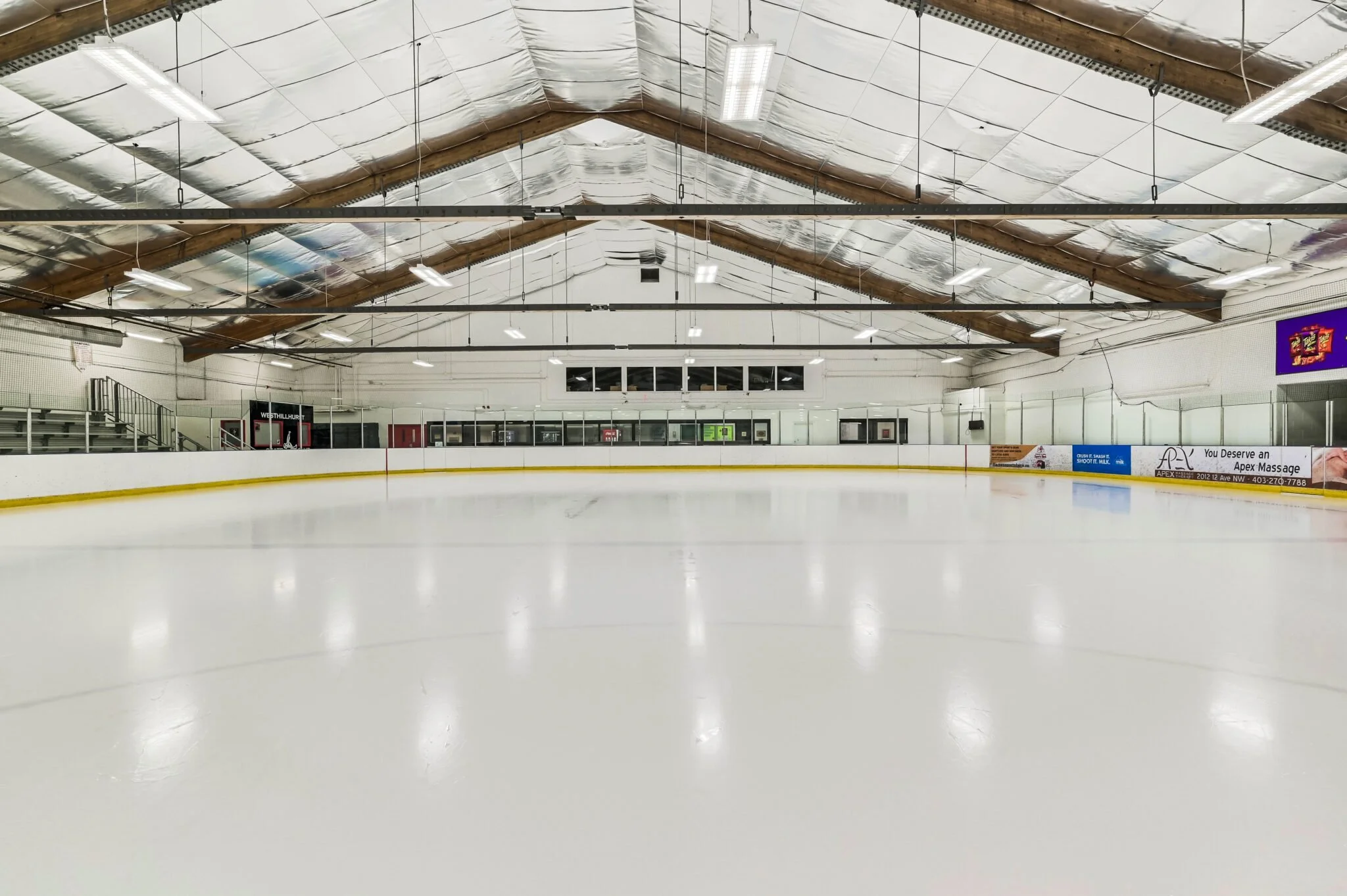 An empty indoor ice hockey rink with white ice surface, surrounded by white boards and clear glass panels. The ceiling has exposed wooden beams and insulation, with several fluorescent lights overhead. Behind the ice, enclosed viewing area with windows and spectators' benches.