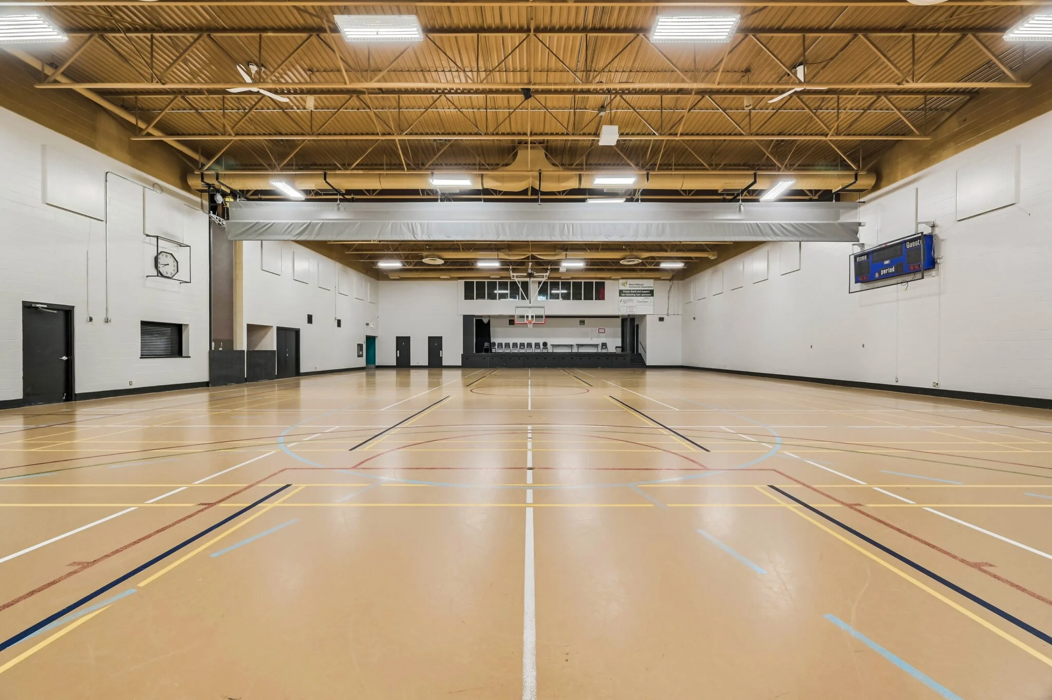 Empty indoor gymnasium with hardwood floor, basketball hoop at the far end, scoreboard on the right wall, and ceiling lights, ready for sports or events.