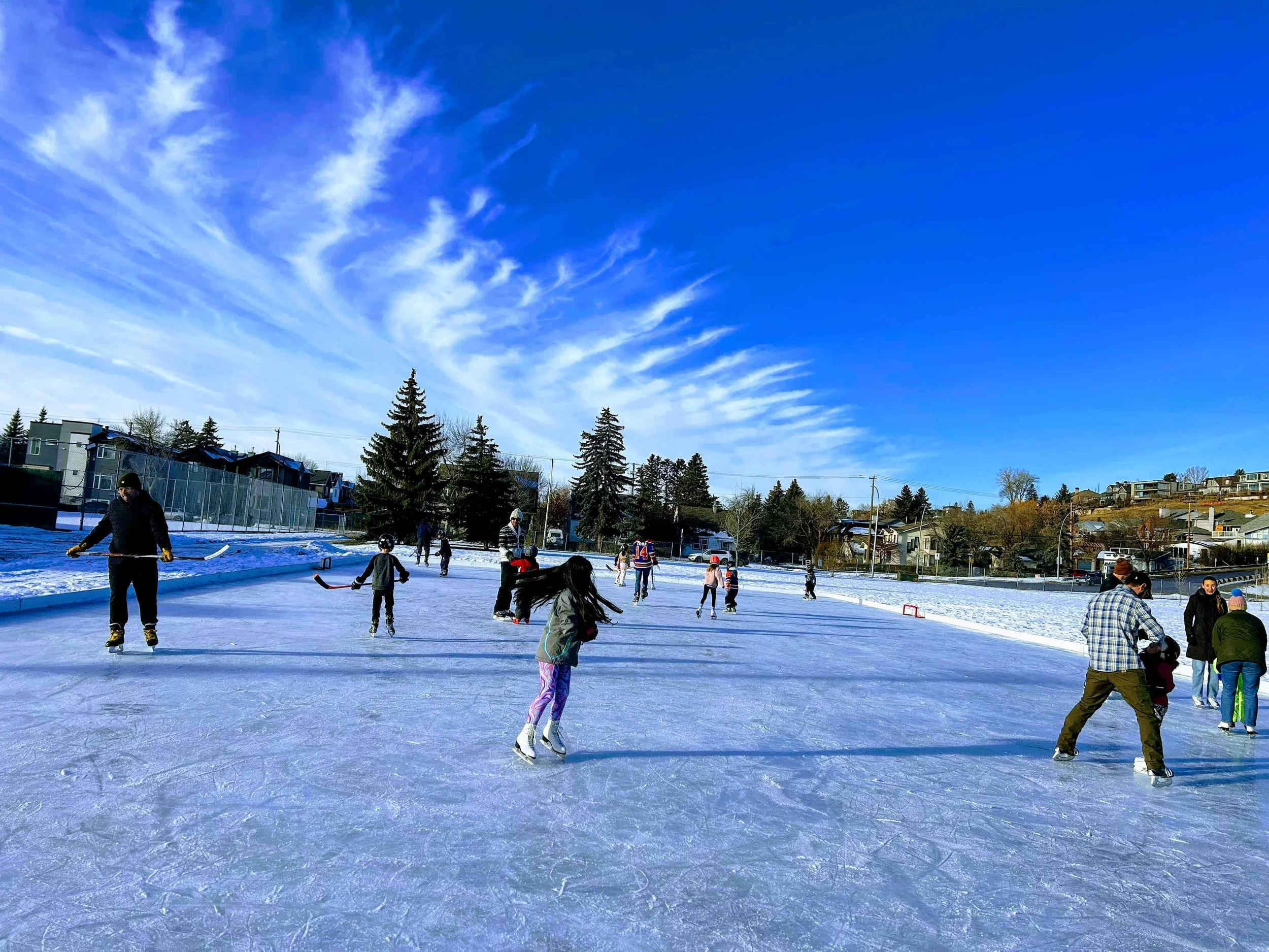 An outdoor ice rink is surrounded by a snowy field. A young girl in purple pants and a green parka is mid twirl in her figure skates. Behind her, there are many small children playing hockey.