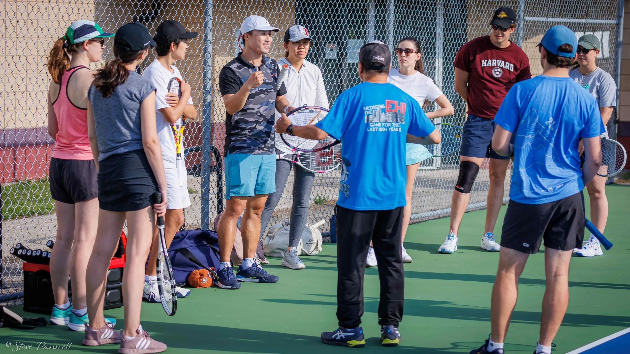 A tennis coach stands on an outdoor court and gives a thumbs up. His students are standing around him in a semi circle. They are holding their racquets.