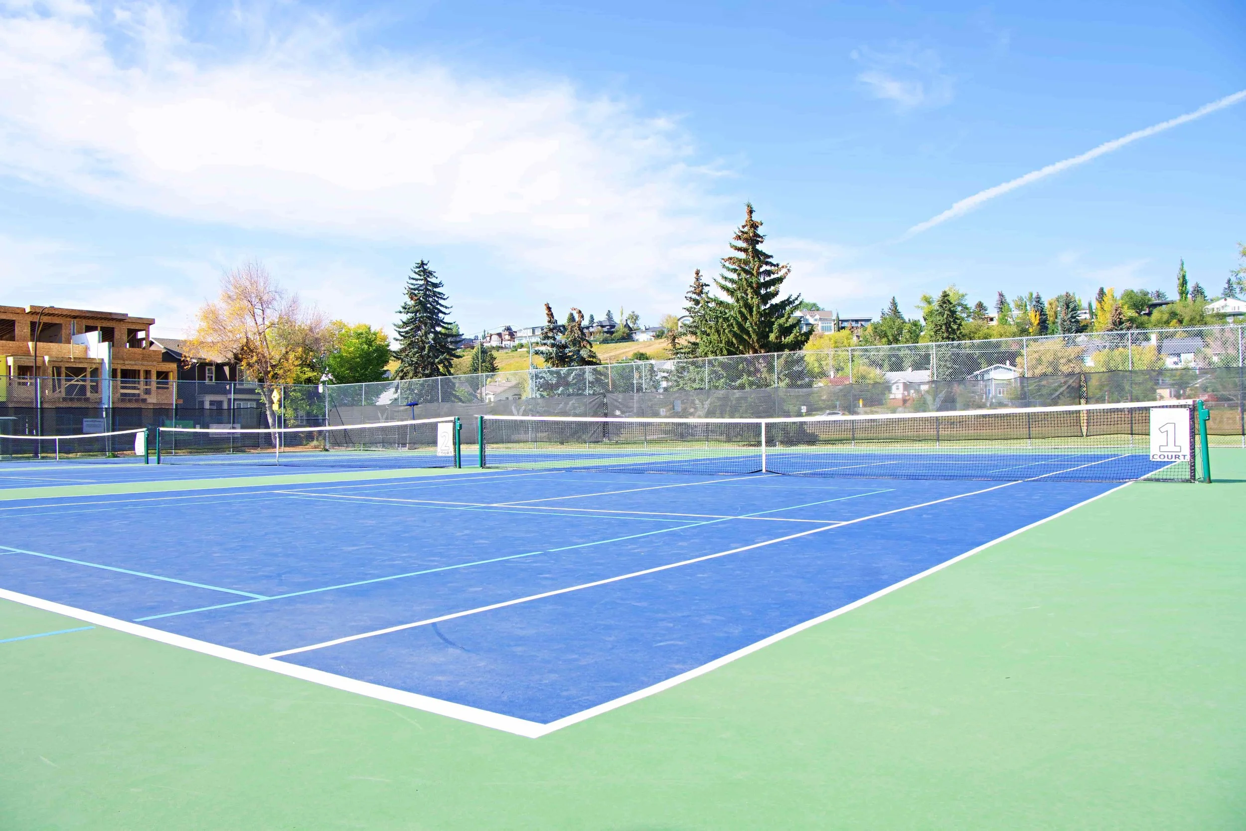 A wide shot of three tennis courts on a sunny day. The courts are green and blue with white lines. In the background there are green trees, a field, and houses