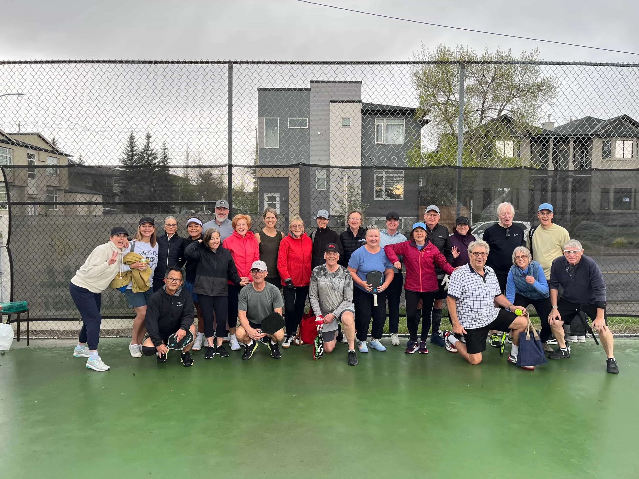 A group of pickleball players of various ages huddle for a group photo on a rainy outdoor court. Behind them is a chainlink fence and houses and trees can be seen in the distance.