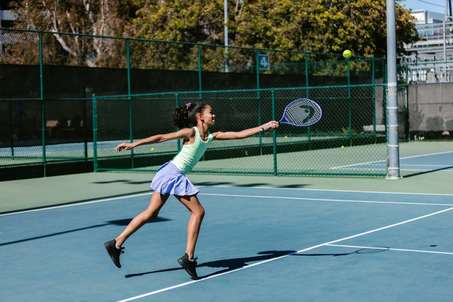 A young girl in a green tank top and a blue skirt playing tennis on an outdoor court, reaching to hit a tennis ball.