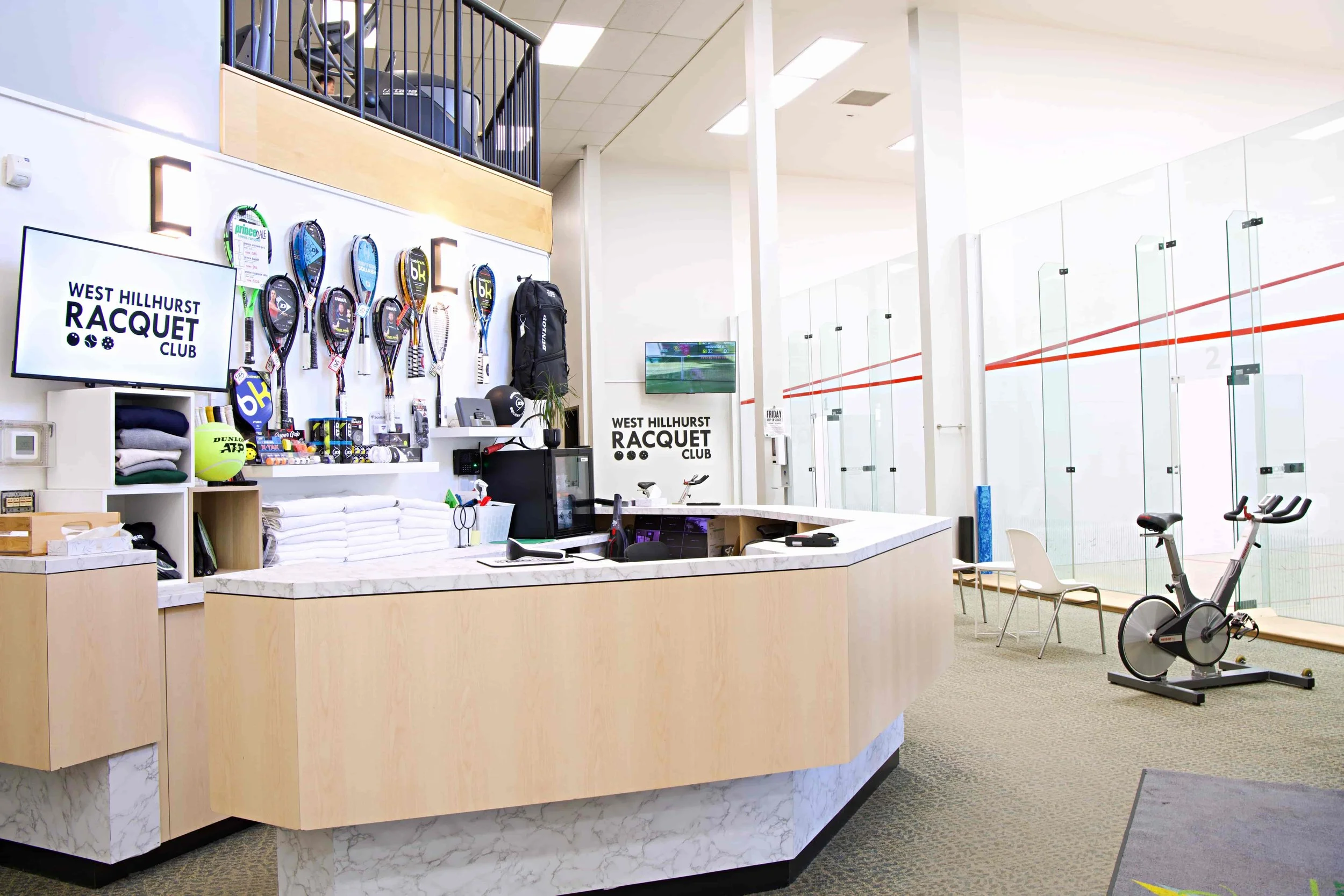 A large, wrap around desk made of light wood and white and grey marble sits in the foreground. On the back wall, there is a TV that reads "West Hillhurst Racquet Club" and several squash racquets hung up.