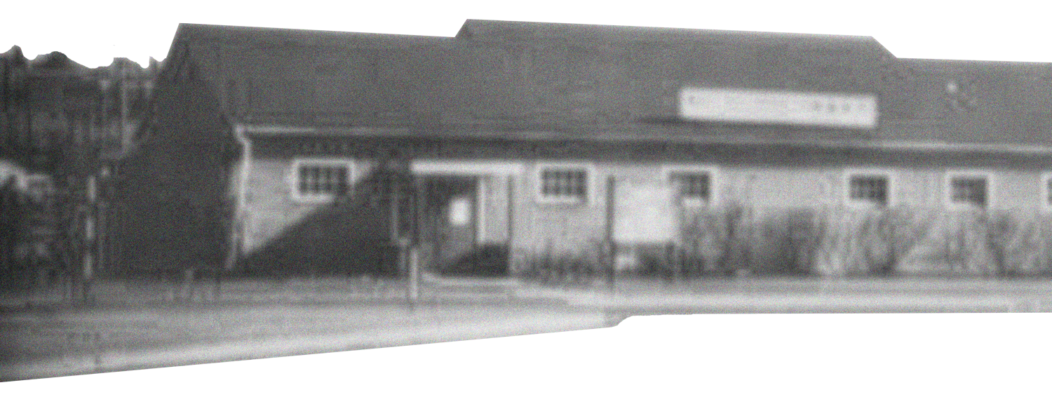 A black-and-white photo of a building with a sloped roof, multiple small windows, and a sign on the front. There is a grassy area with trees and a sidewalk in front of the building.