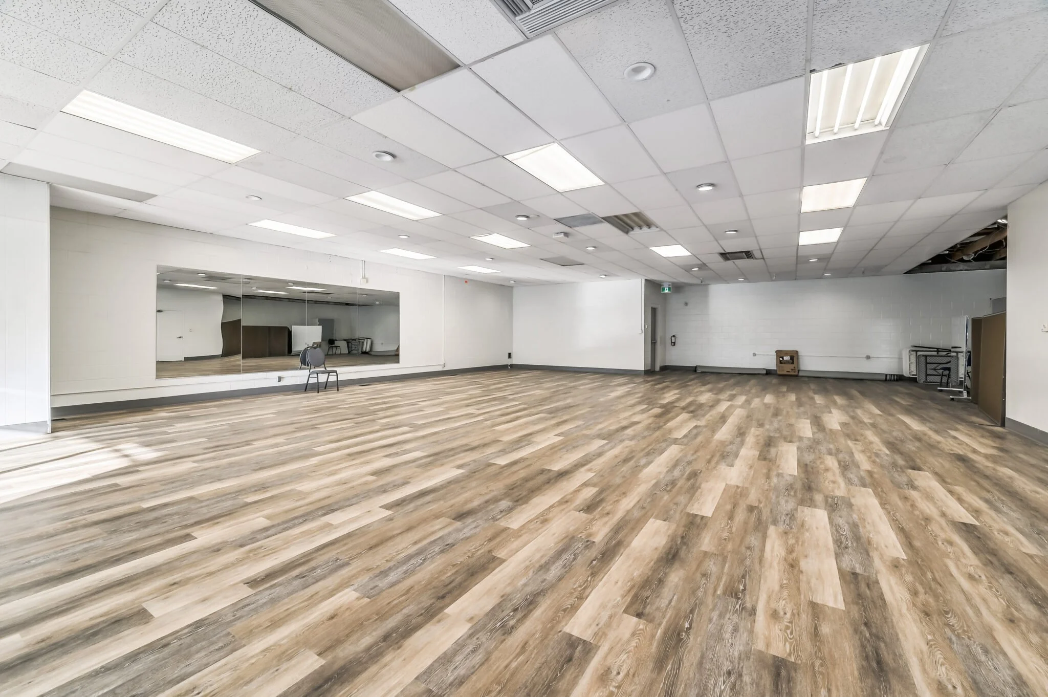 Empty dance or exercise studio with wood-like flooring, white walls, a large mirror on one wall, and fluorescent ceiling lights.