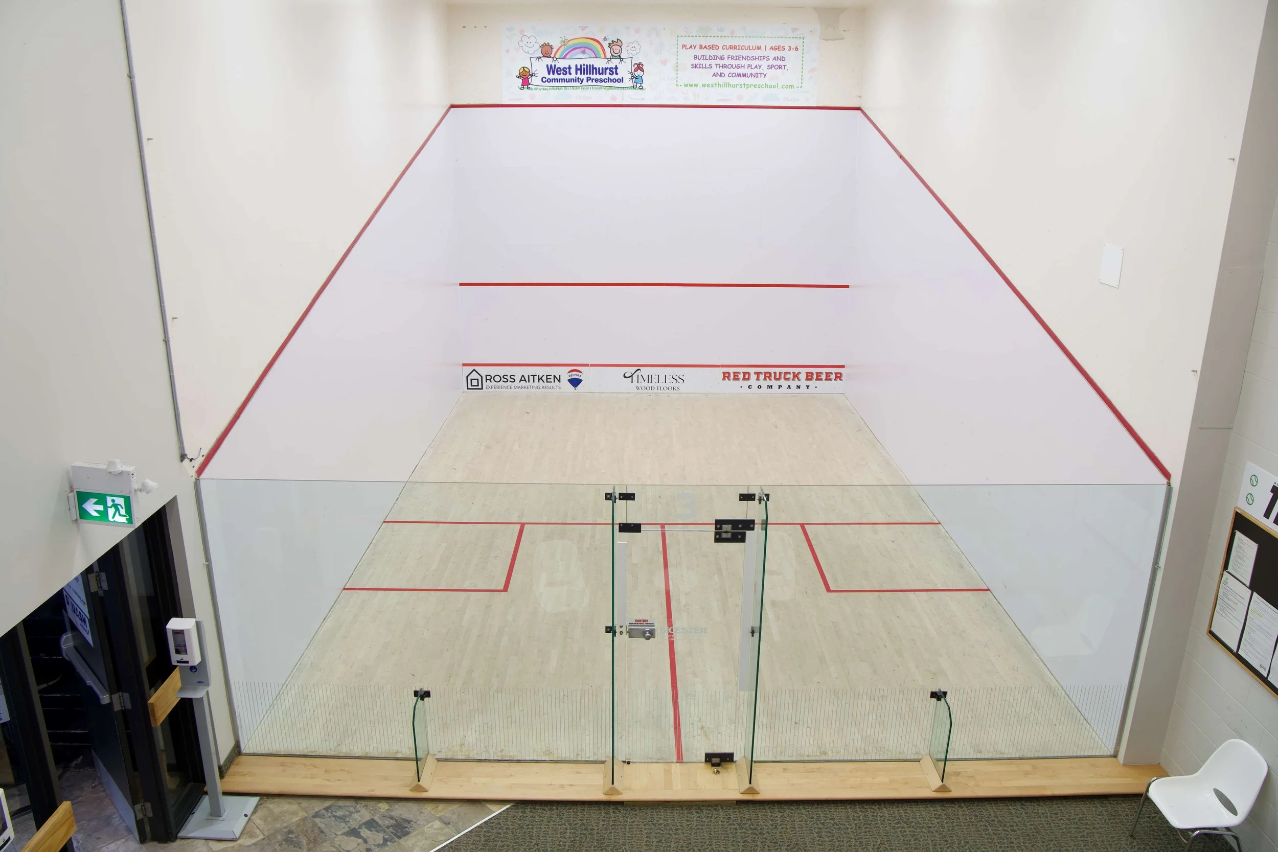 An aerial view of a squash court with light wooden floors, white walls, red lines, and glass doors. There are several rectangular advertisements located on the wall, including three small ones along the bottom and one large one at the top.