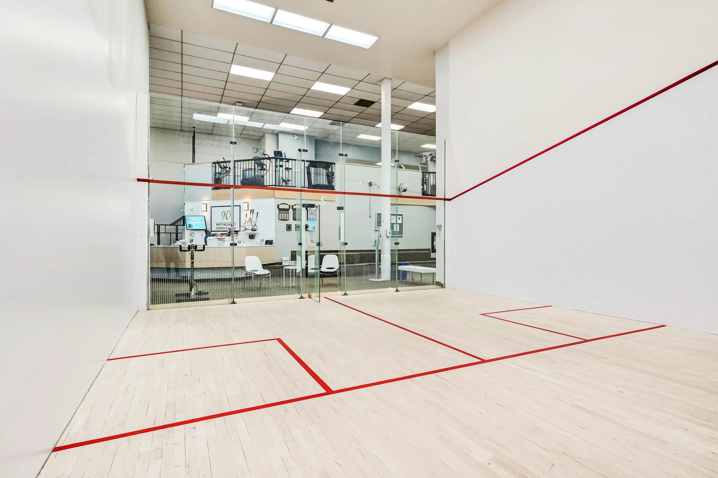 Indoor squash court with light wooden floor and red boundary lines, enclosed by glass walls, overlooking a fitness center with exercise equipment and seating area.