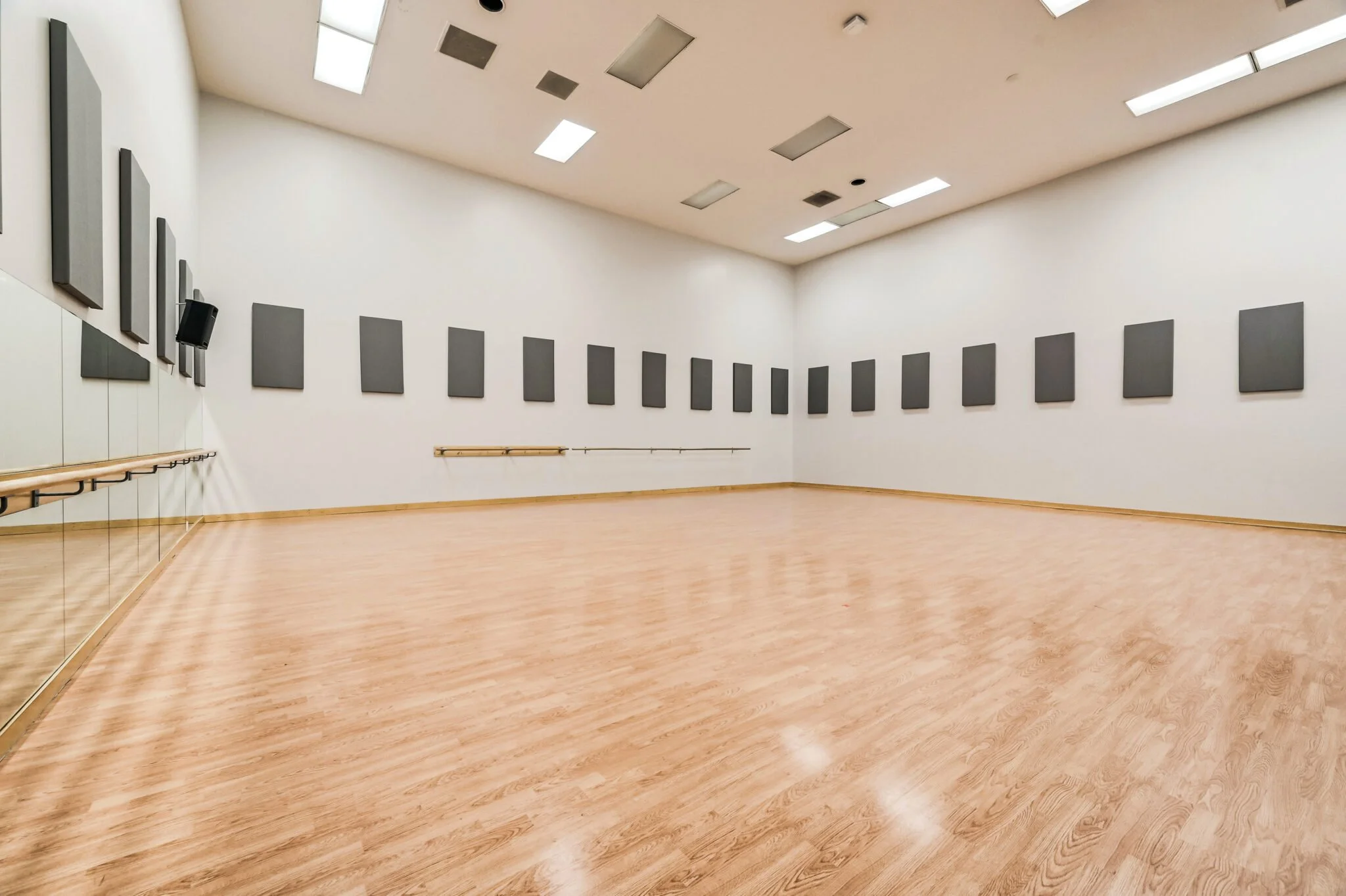 Empty dance studio with wooden floor, black sound panels on white walls, mirrors on left side, and ceiling lights.