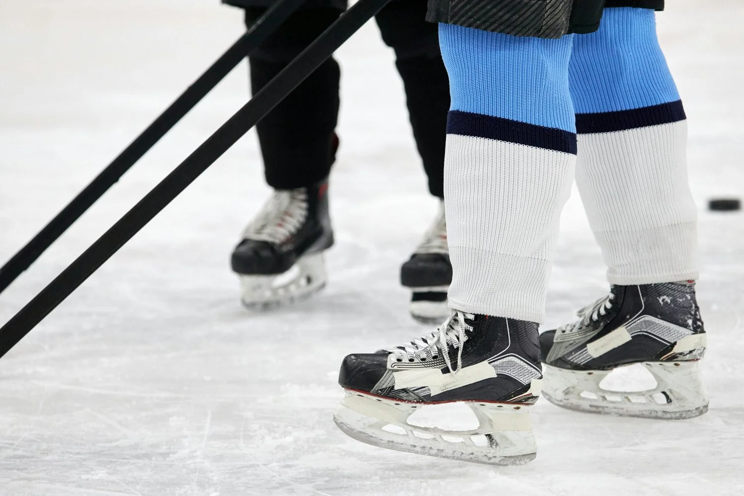 Close-up of ice hockey skates and hockey socks on players standing on ice rink, with hockey sticks visible in the foreground.
