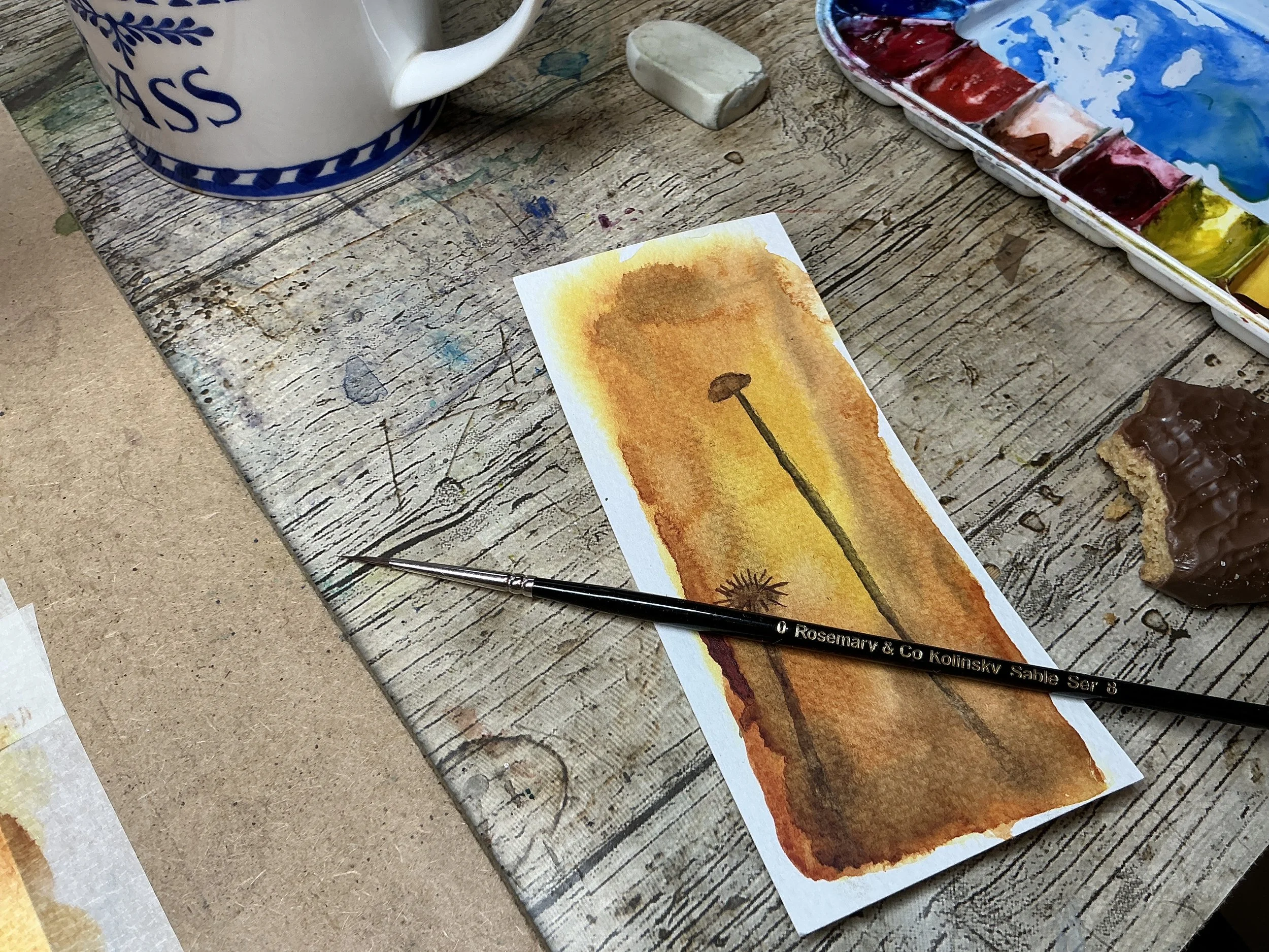 Watercolor painting of a dandelion seed head with a yellow-orange background, on a wooden table with watercolor supplies and a mug nearby.