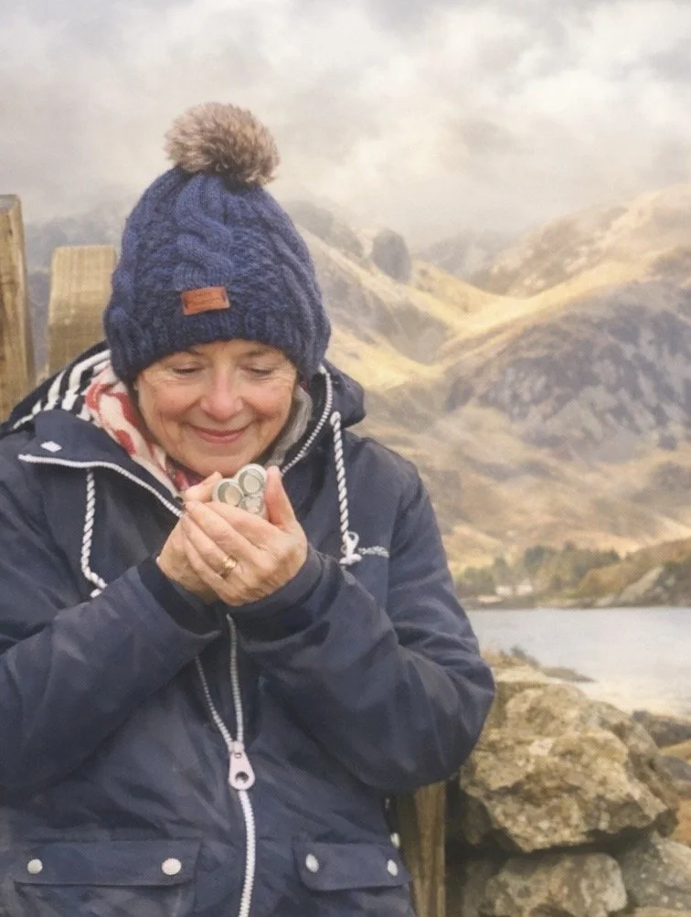 A smiling woman wearing a dark blue winter jacket and a blue knit beanie with a pompom, holding three small silver objects, standing outdoors near a body of water with mountains in the background.