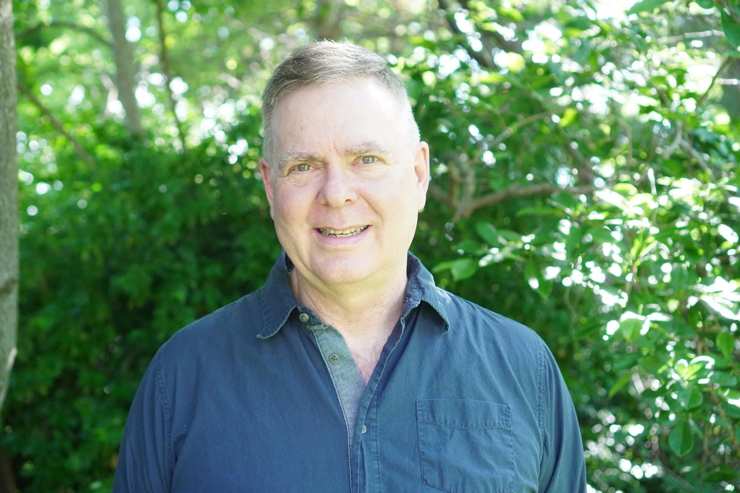 A headshot of a middle-aged man with a background of greenery in a blue buttoned shirt.