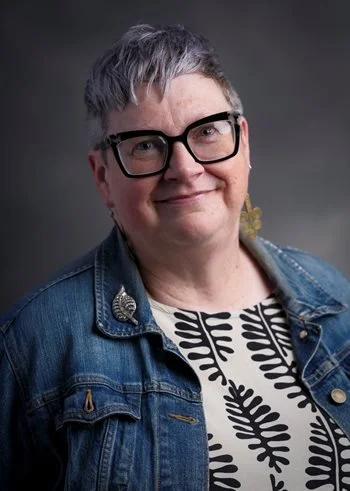Headshot of Lynn R. Huber in front of a gray background. Lynn is wearing black frame glasses, a jean jacket, and a white and black patterned top. Lynn has gray hair in a pixie cut.