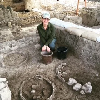Photo of Cynthia Shafer Elliott at a dig site, surrounded by dirt and artifacts. Cynthia is in a squat position and is smiling, wearing a baseball cap, olive top, and blue jeans.
