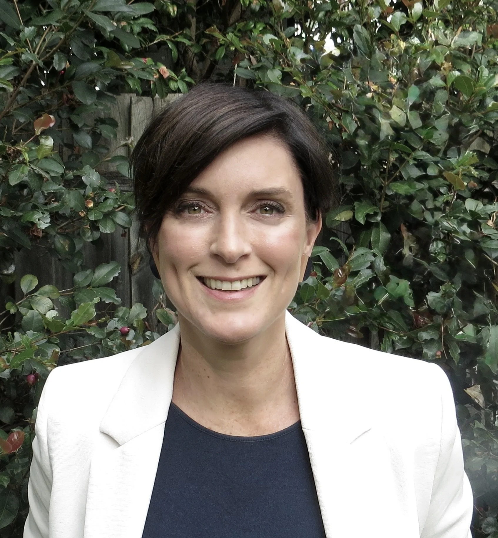 A photo of Robyn Whitaker with greenery in the background. She is wearing a white jacket with a dark blue shirt underneath. She has short, dark hair. She is smiling at the camera.