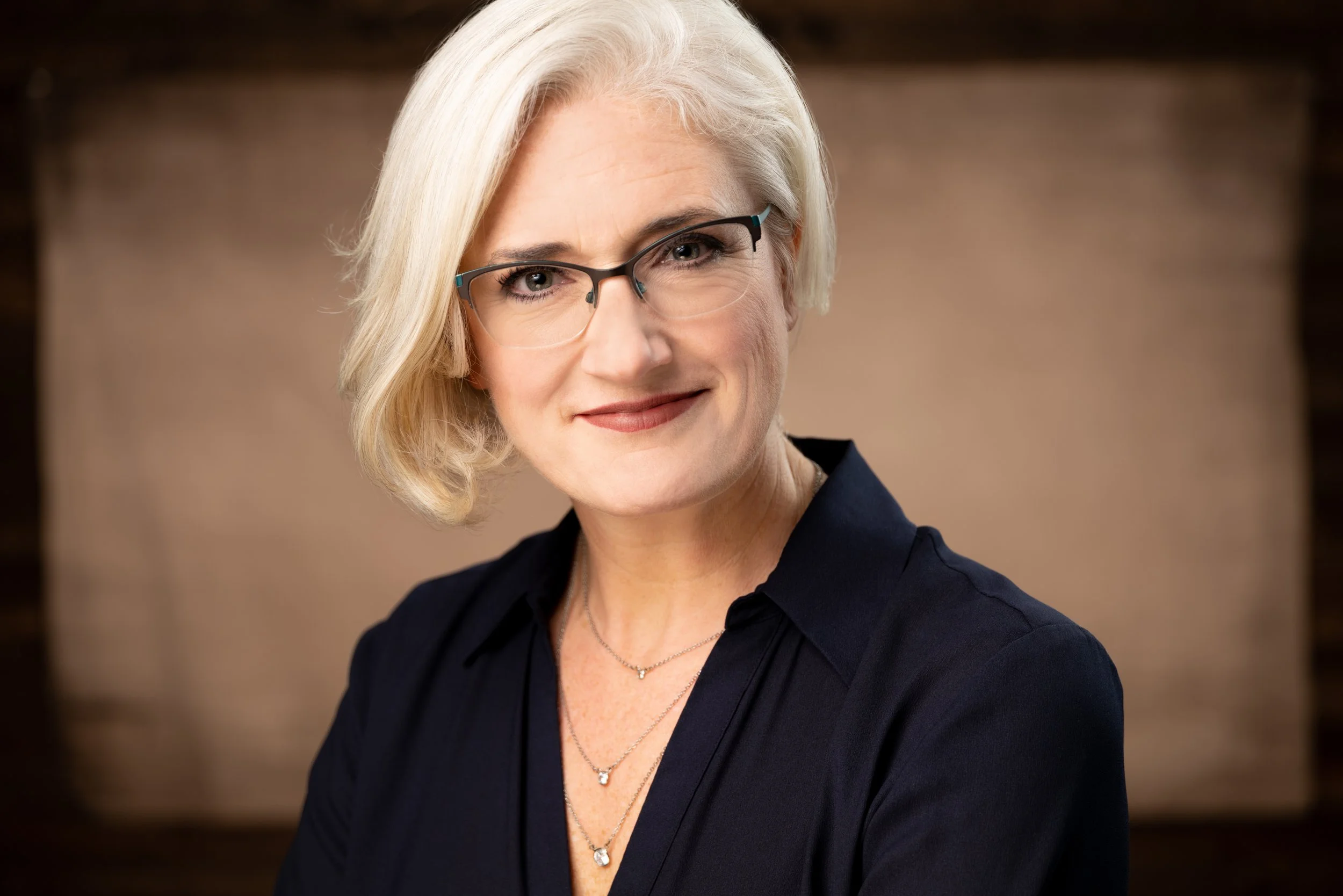 Headshot of Dr Jennifer G Bird with a brown background. She has a black shirt, necklaces, and glasses. Her hair is short and light colored.