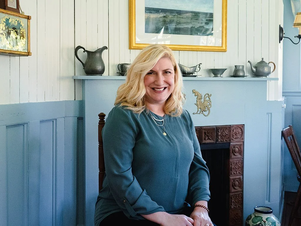 Headshot of Anna Sieges-Beal in front of a wall with a blue fireplace and white shiplap. Anna has blonde hair and is wearing a blue top.