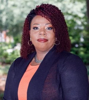 Headshot of Angela N. Parker with outdoor background including green tree leaves. Angela is wearing a blue cardigan over a coral top.