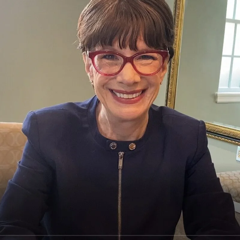 Headshot of Amy-Jill Levine wearing a collarless zip up blazer and red cat-eye glasses sitting on a tan chair with beige walls and a mirror in the background.