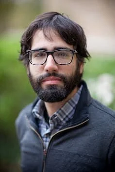 Headshot of Roberto de La Noval. Roberto is wearing black frame eyeglasses and is in front of greenery and a brick building. He has dark mid length hair and a beard.