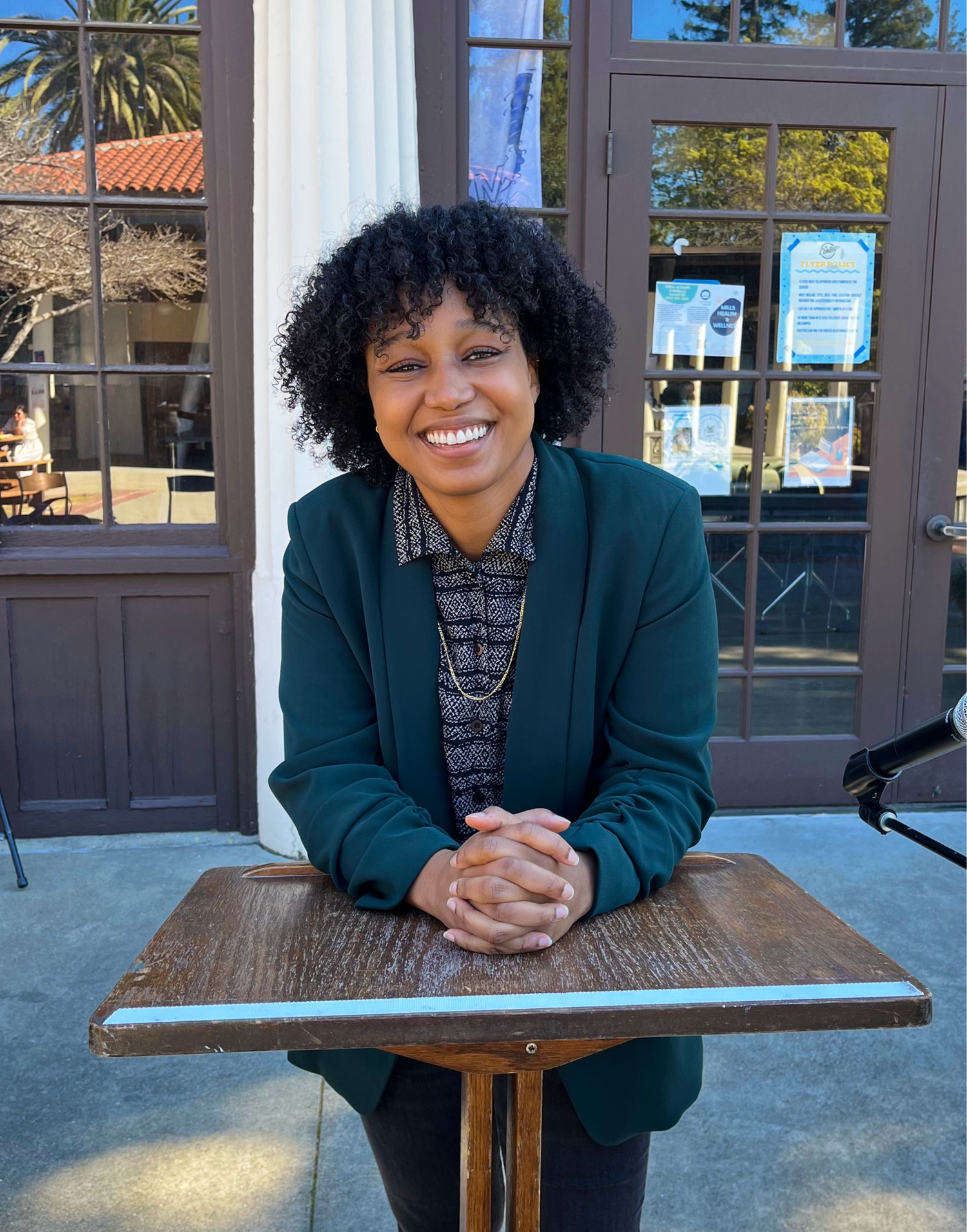 A photo of a woman smiling while leaning on an easel. She is wearing a blue jacket over a black and white dress shirt.