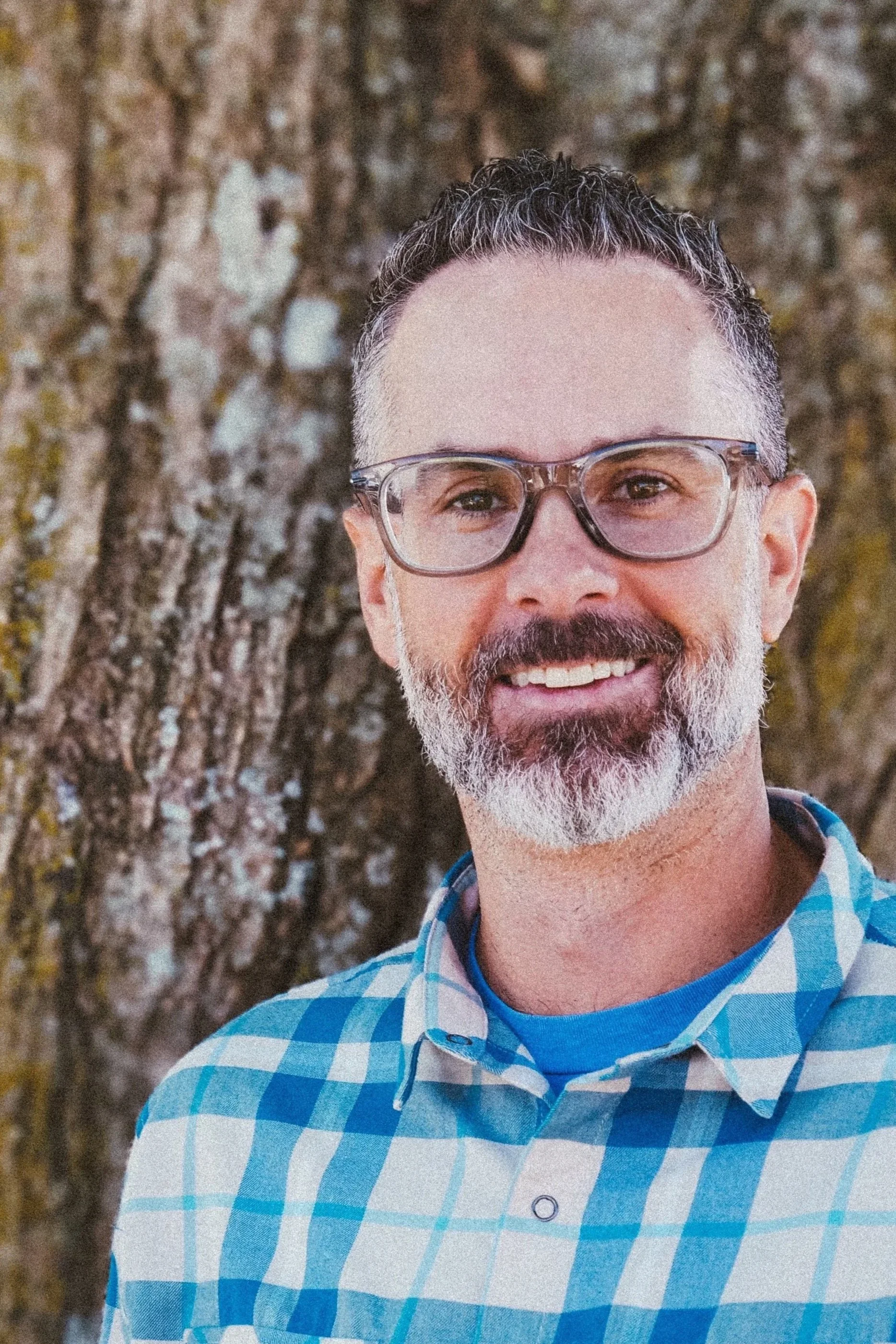 Headshot of Joshua James in front of a tree. He is wearing a blue and white plaid button down shirt.