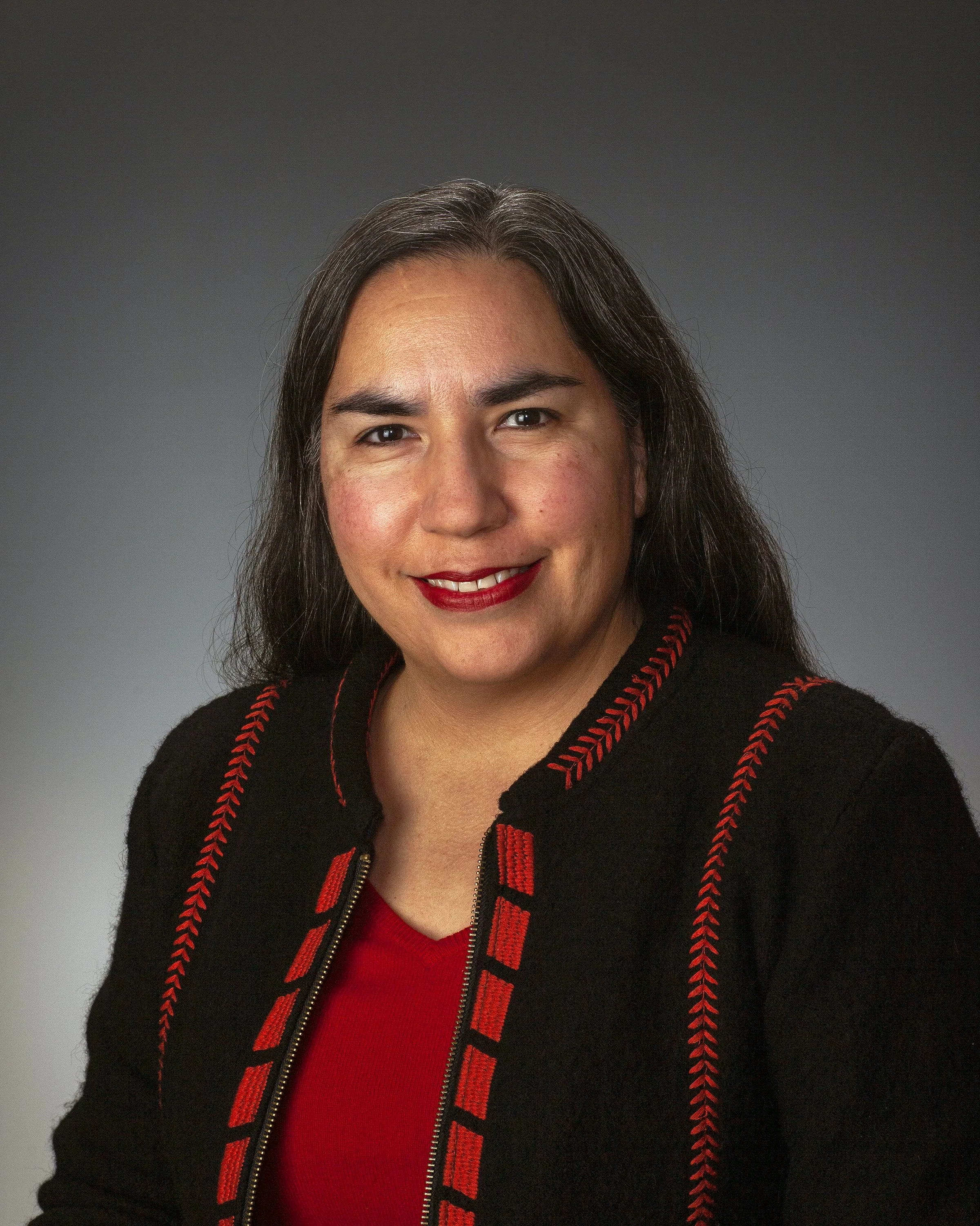 Headshot of Sarah Augustine in front of a gray background. Sarah is wearing a black and red jacket with indigenous weaving patterns over a red top.