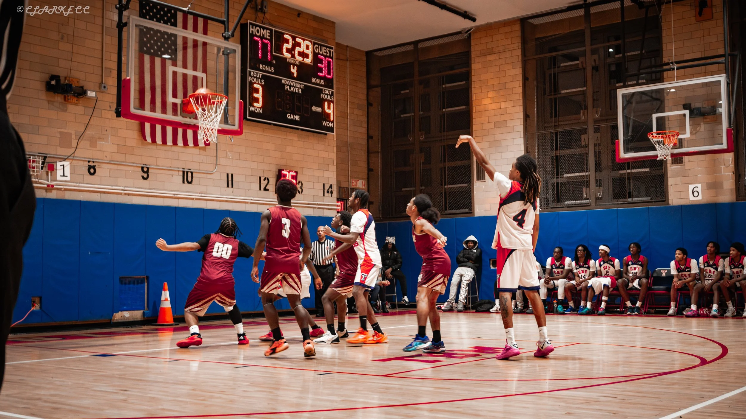 A basketball on an indoor court with a scoreboard showing 77-30, with players in red and white uniforms and spectators sitting on the side