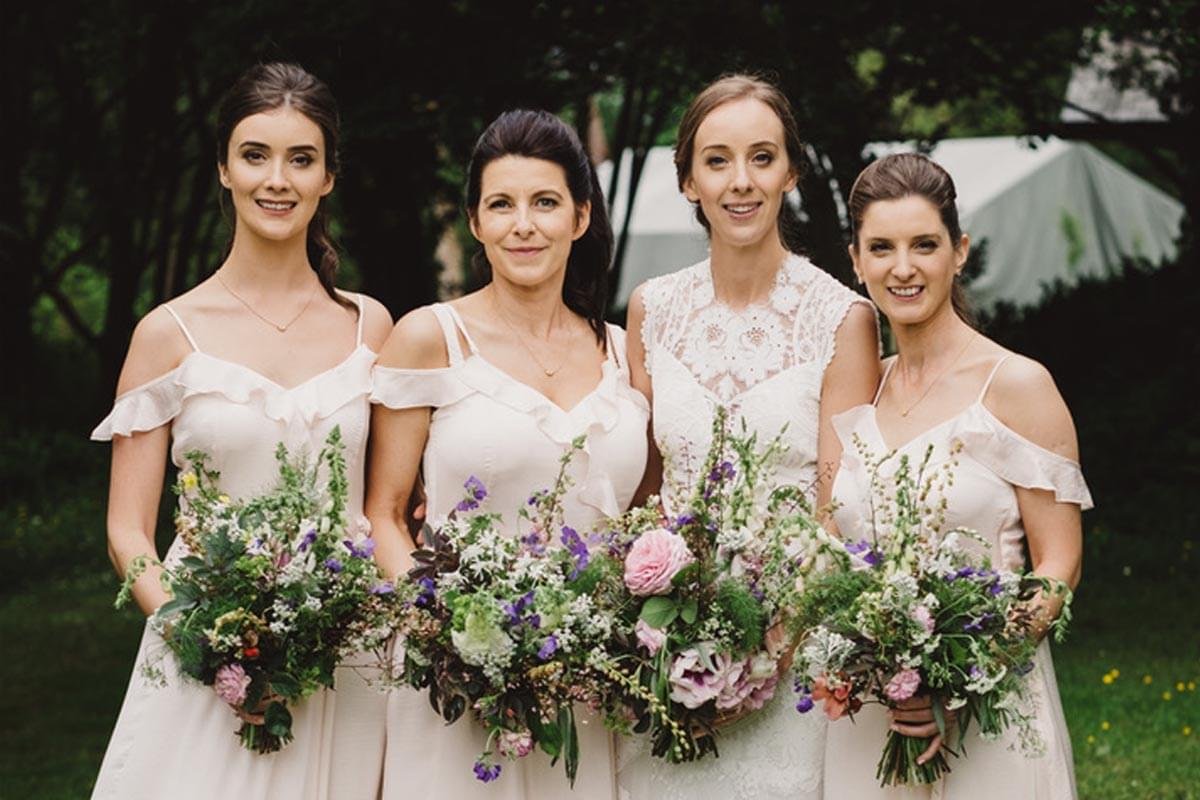 Four women in white dresses holding floral bouquets standing outdoors in a garden setting.