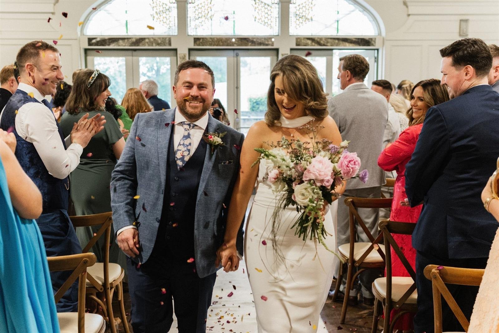 A newly married couple walking down the aisle holding hands in a wedding ceremony, surrounded by friends and family, with confetti falling around them.