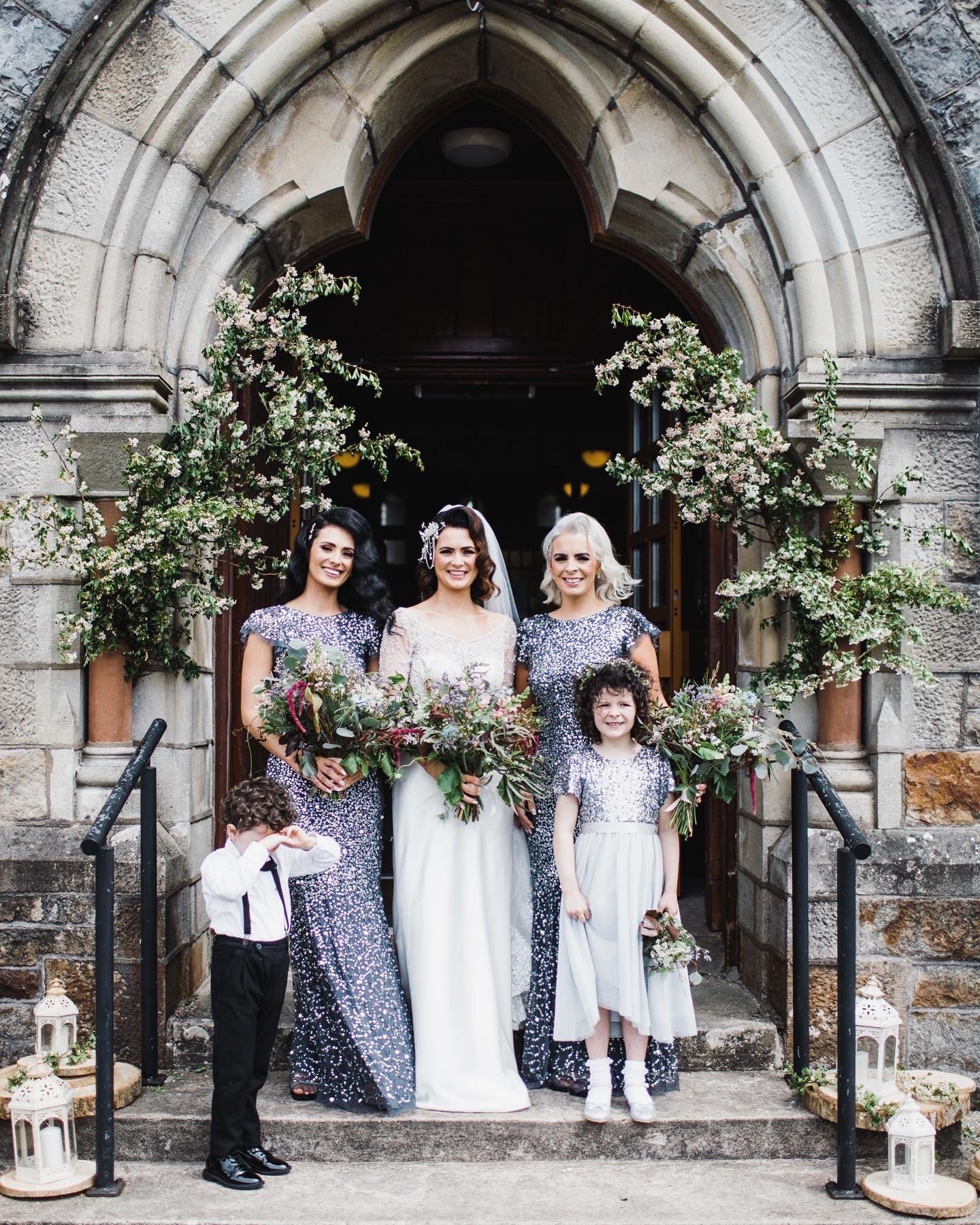 Group of women and children standing on church step, celebrating wedding, with floral decoration. Woman in wedding dress, holding bouquet, in center. Two women and two children around her, all holding flowers.