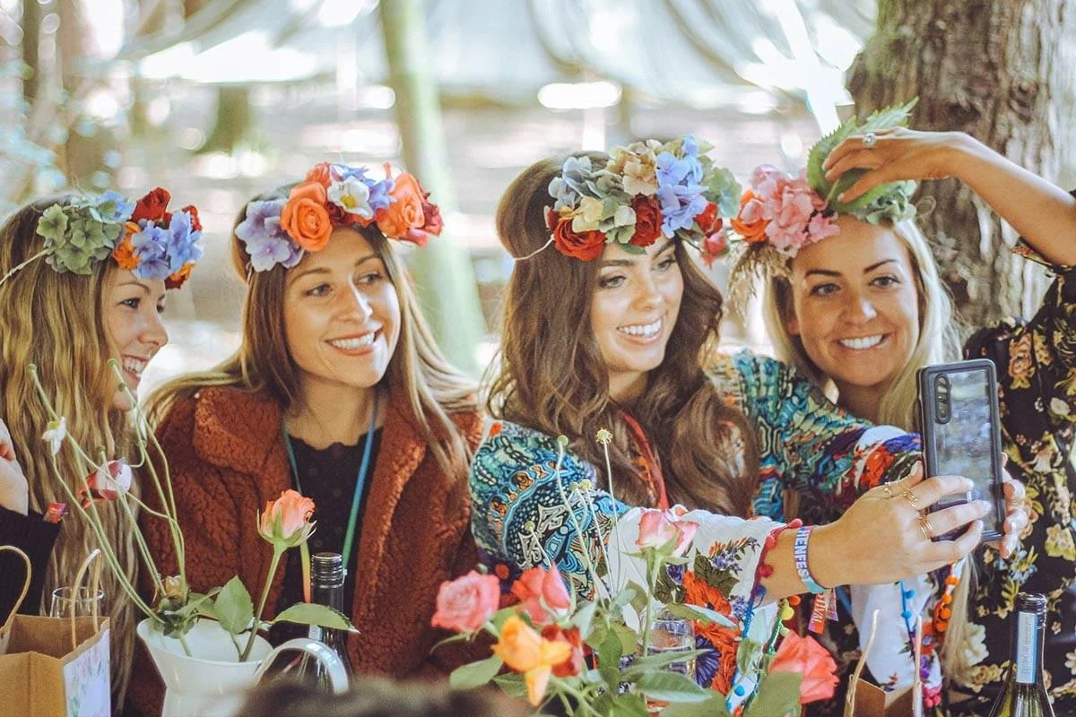 Four women wearing flower crowns take a selfie together during a celebration or gathering outdoors.