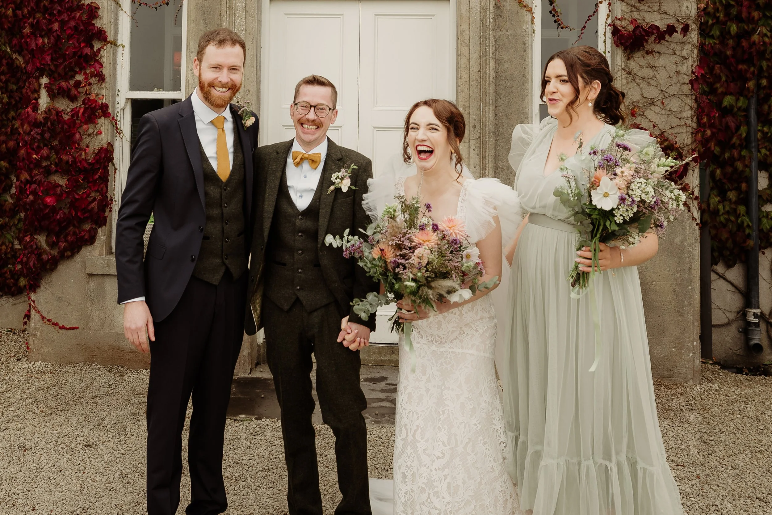 Group of five people at a wedding, two men and three women, standing outside a building with ivy-covered walls. The bride is holding a bouquet and laughing. The woman on the far right is also holding a bouquet. The two men are wearing suits with vest