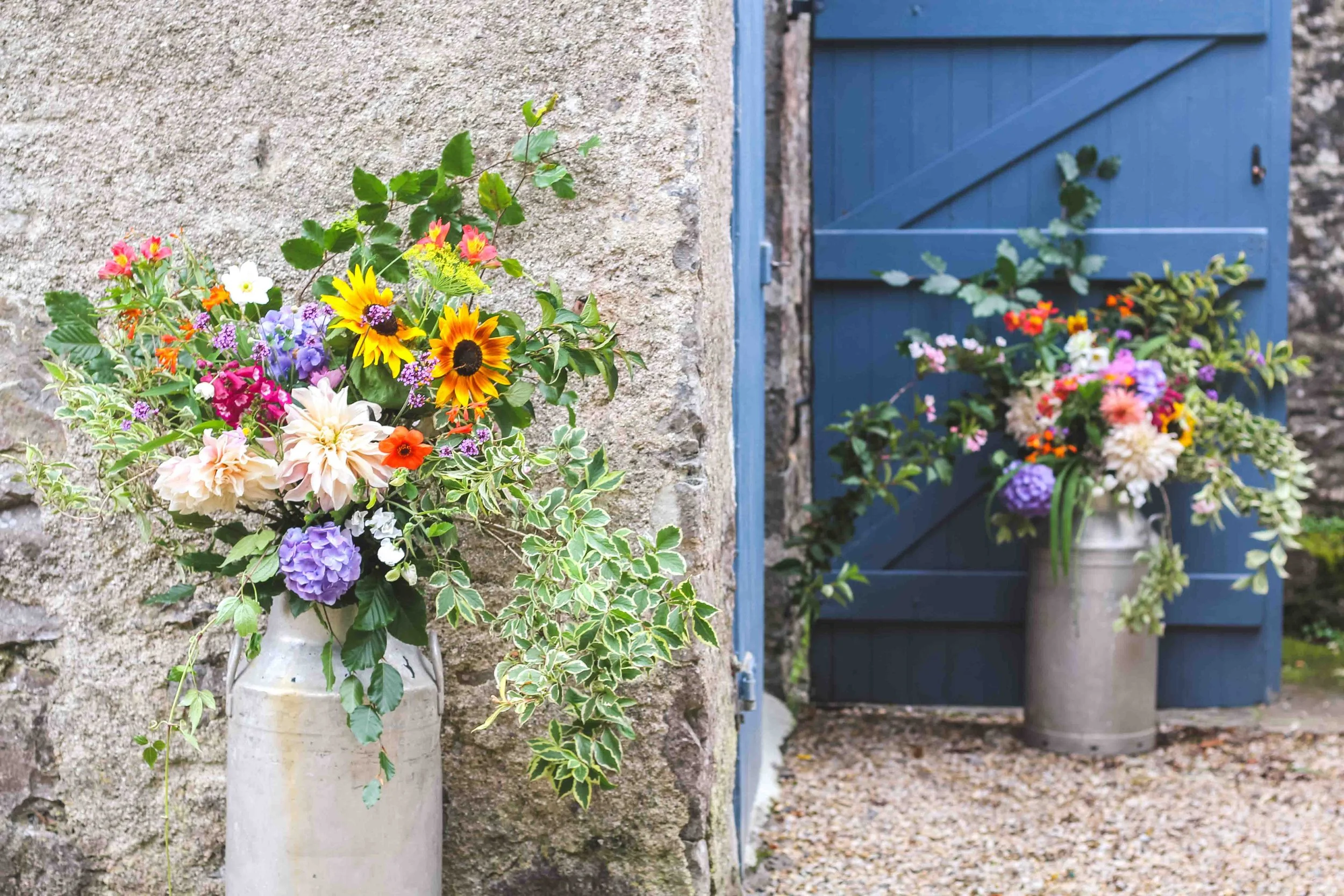 A large vase with colorful flowers, including sunflowers and dahlias, placed on a stone ledge beside a blue wooden door and wall, with another large vase of flowers outside the door.