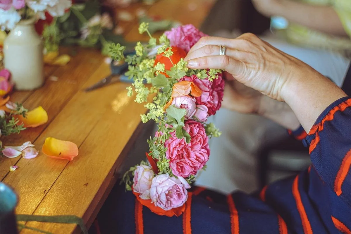 Person arranging a flower crown with pink, red, and orange flowers on a wooden table decorated with flower petals.