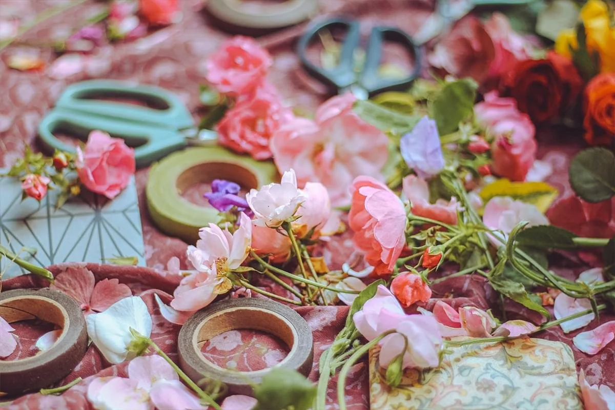 A variety of floral supplies including colorful flowers, floral scissors, tape, and floral patterned paper scattered on a pink fabric surface.