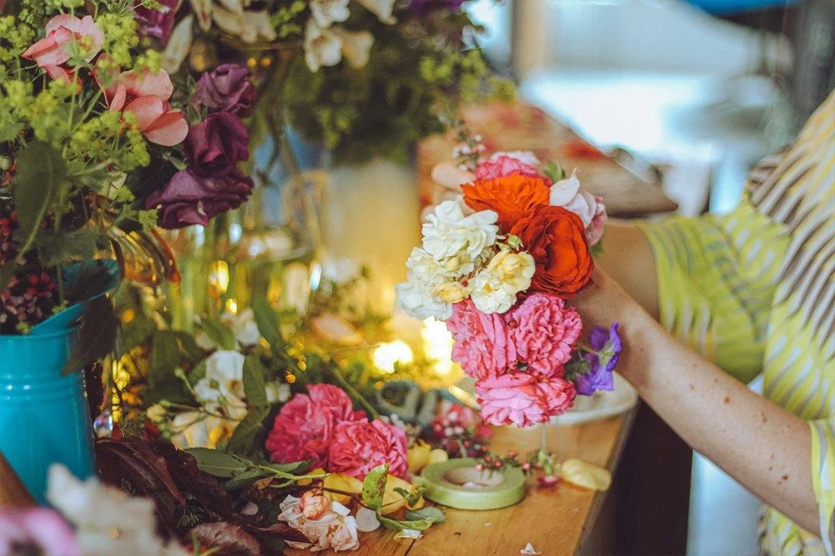 A person arranging colorful flowers into a bouquet on a wooden table, with a variety of flowers and greenery nearby, in a well-lit room.