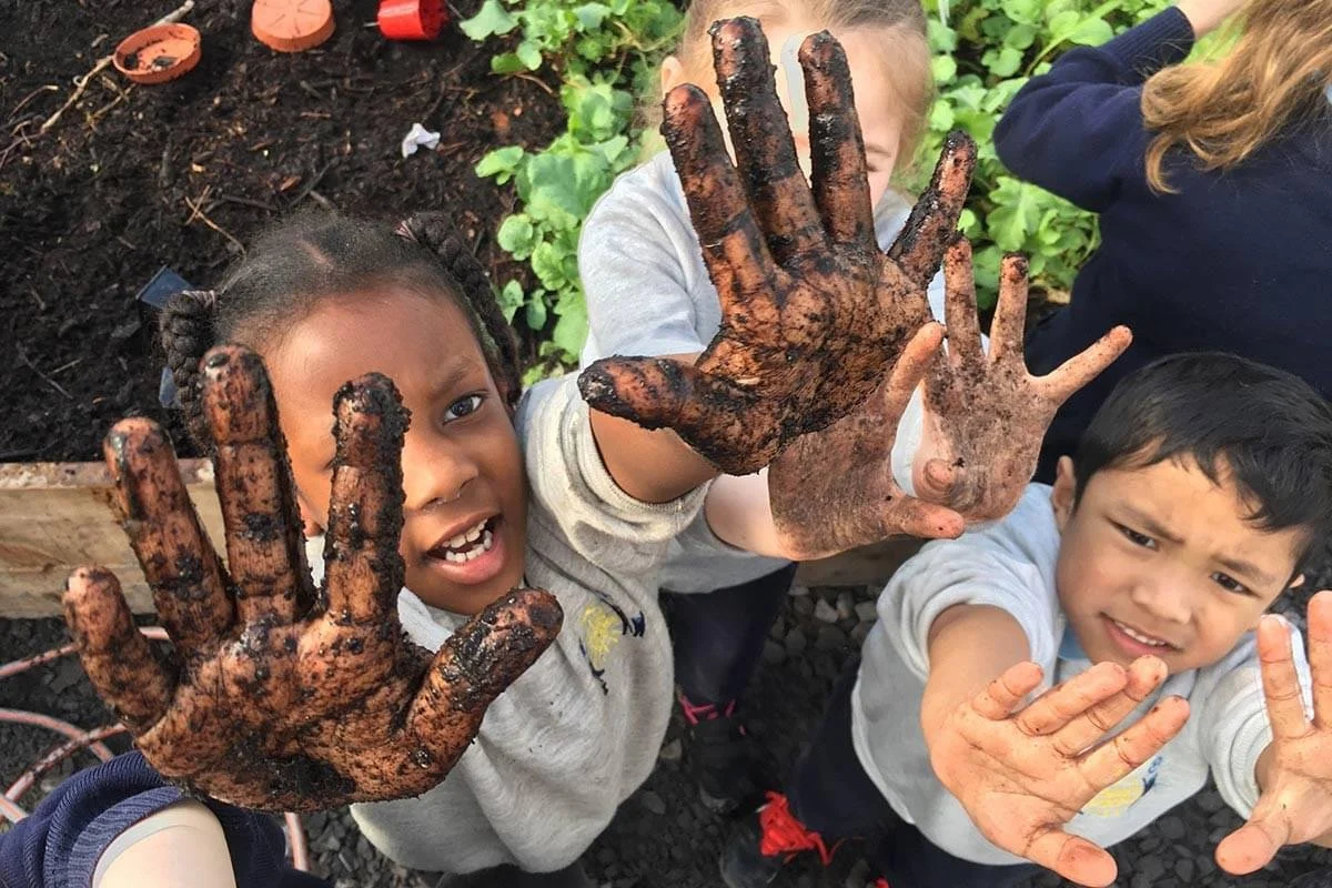 Two children with dirty hands showing front of their hands, smiling and looking at the camera in a garden area.